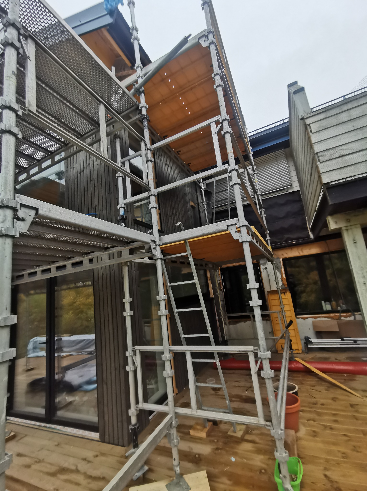 Montaż elewacji domu z ciemnego drewna, widoczne rusztowanie i elementy konstrukcyjne.