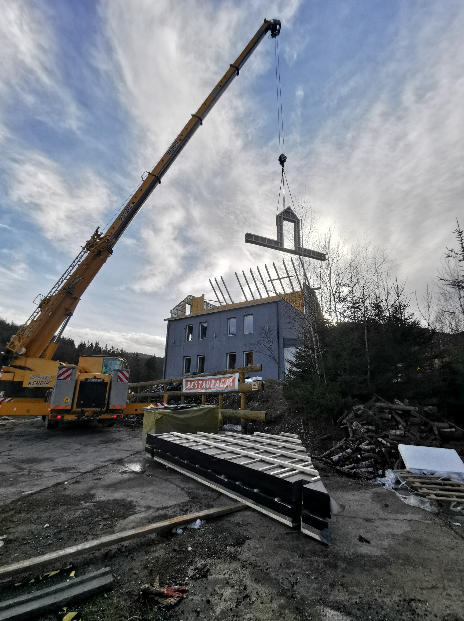 Montaż elementu szkieletowego domu z użyciem dźwigu na tle pochmurnego nieba i częściowo wykończonego budynku.