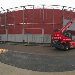 Szeroki kadr na budowę hali z czerwonymi panelami, gdzie pracownicy na podnośnikach montują szare płyty warstwowe, a na pierwszym planie leżą palety z materiałami.