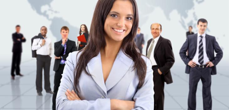 Uśmiechnięta kobieta w jasnym żakiecie na tle grupy biznesmenów i mapy świata, sugerująca międzynarodowy zespół lub globalny zasięg.