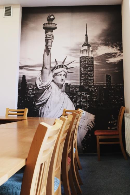 Sala lekcyjna z długim stołem i krzesłami, na ścianie fototapeta przedstawiająca Statuę Wolności i panoramę Nowego Jorku w czerni i bieli.
