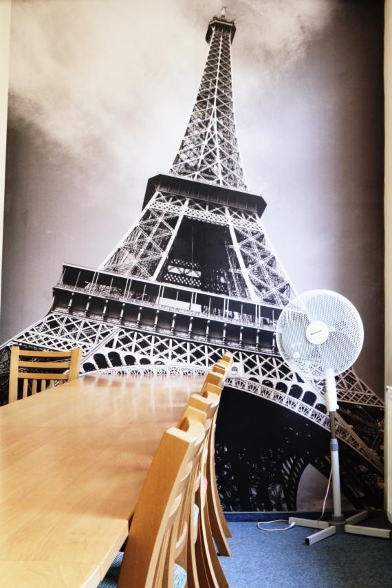 Sala lekcyjna z długim stołem i krzesłami, na ścianie fototapeta z wieżą Eiffla, w rogu stoi włączony wentylator.