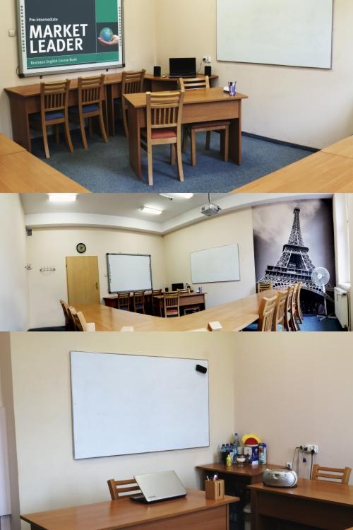 Sala lekcyjna z drewnianymi ławkami i krzesłami, tablicą suchościeralną, plakatem z napisem 'Market Leader' i tapetą z wieżą Eiffla, przygotowana do zajęć językowych.