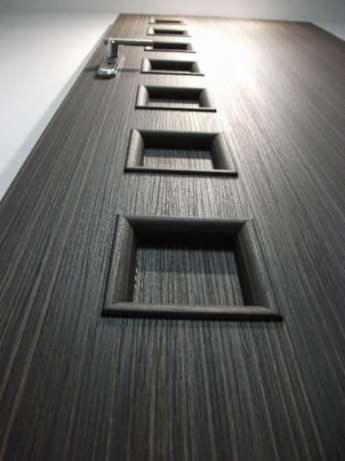 Ciemnoszare drzwi z drewnopodobną fakturą i rzędem kwadratowych wgłębień oraz srebrną klamką, sfotografowane z dołu pod kątem.