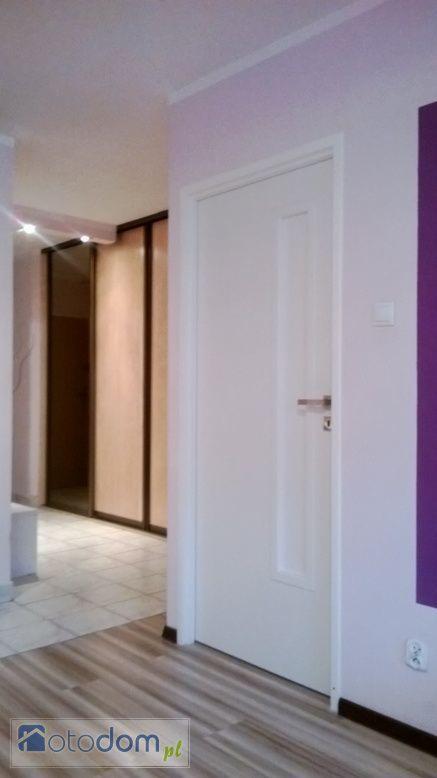 Wnętrze mieszkania po remoncie: białe drzwi wewnętrzne z panelem dekoracyjnym, widok na szafę przesuwną w korytarzu z panelami podłogowymi imitującymi drewno.