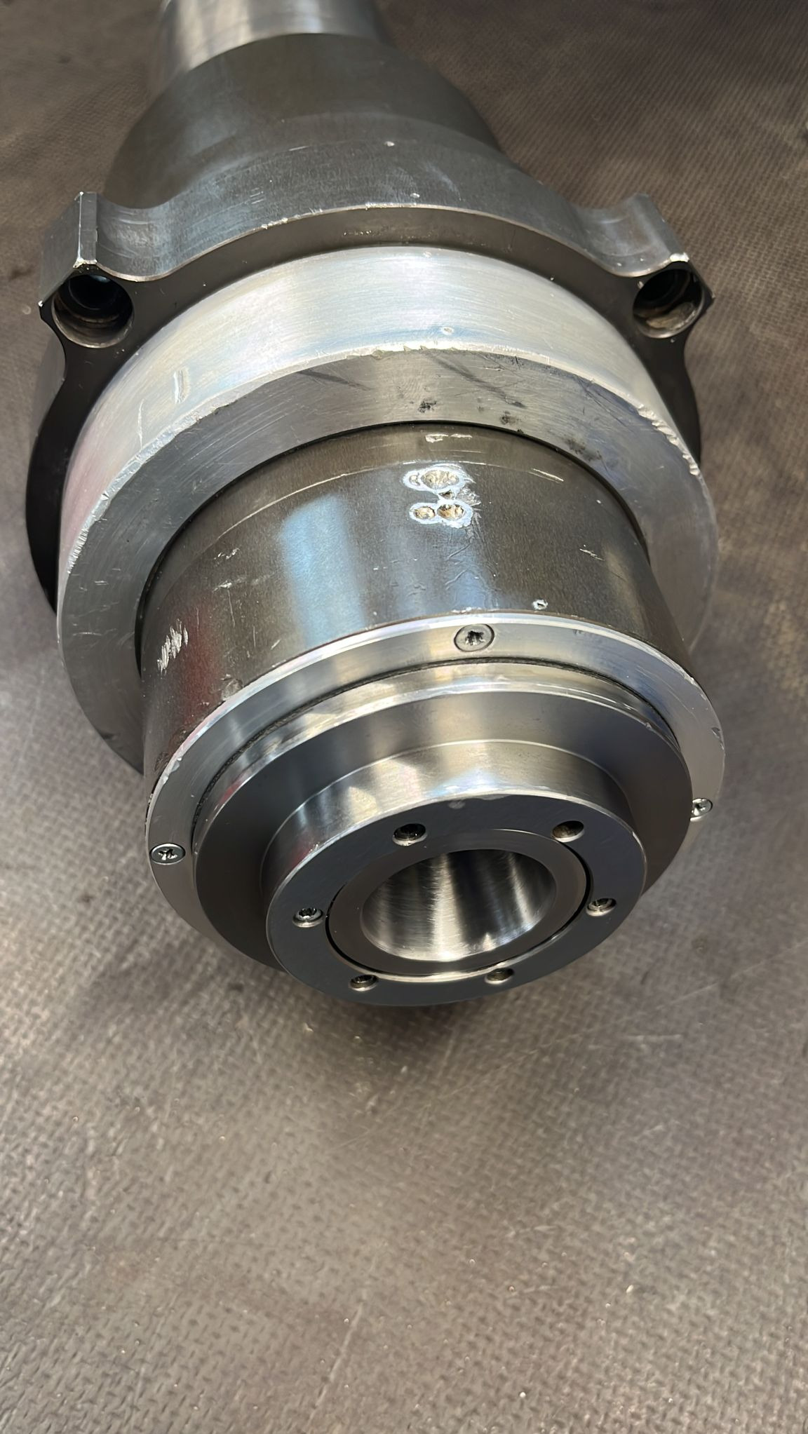 Metalowe wrzeciono tokarskie z widocznymi śladami użytkowania, mocowane śrubami, leżące na szarej powierzchni.