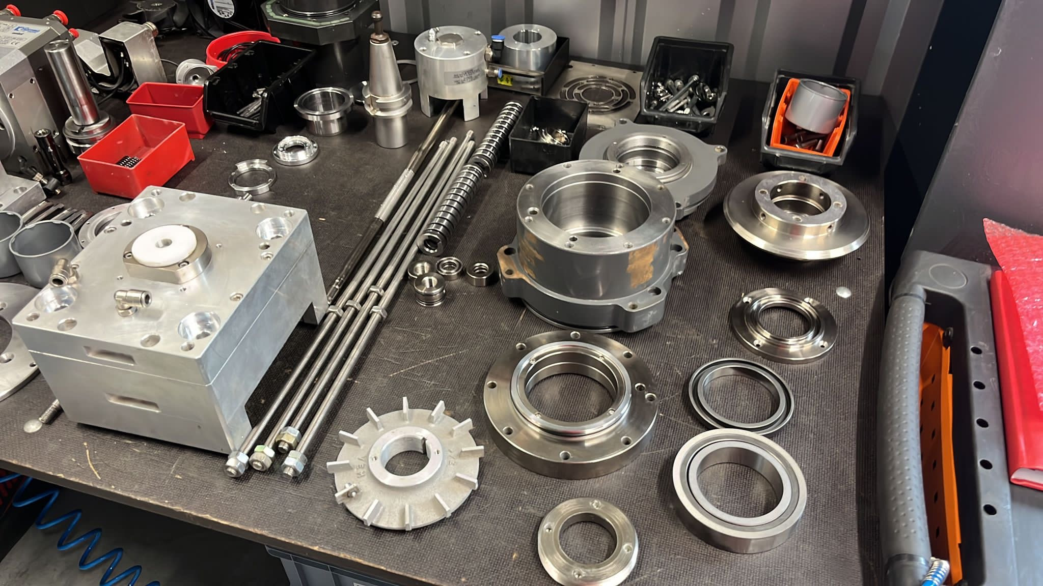 Aluminiowy blok z białym elementem na górze, kilka metalowych pierścieni, łożysk i prętów gwintowanych leżących na czarnym stole warsztatowym.