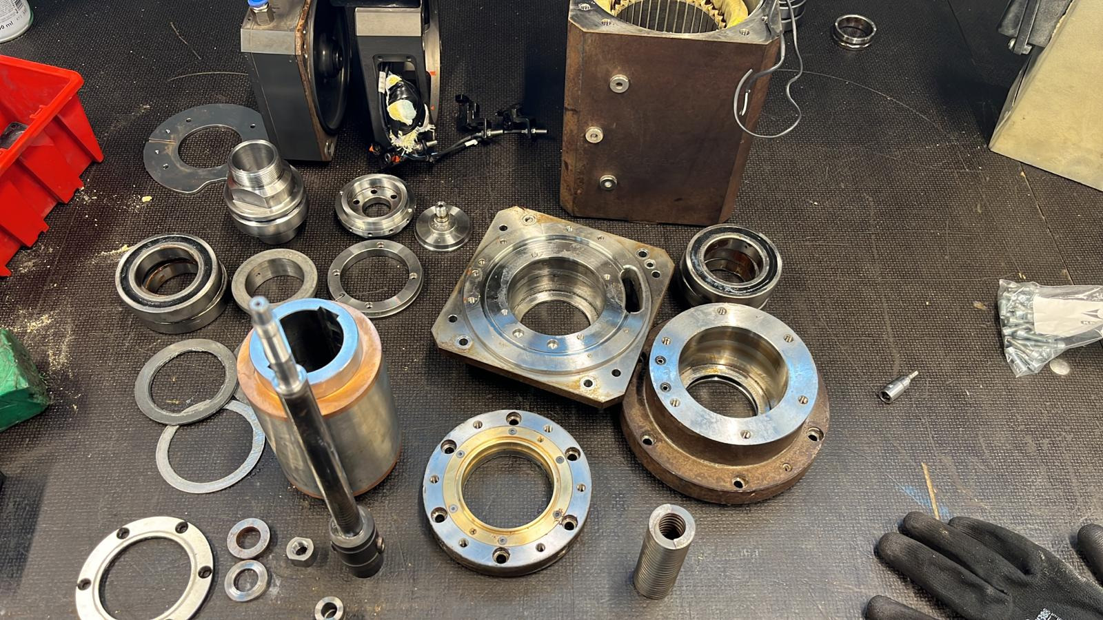 Rozłożone części metalowe, w tym łożyska i tuleje, prawdopodobnie elementu maszyny, leżące na ciemnym blacie roboczym, obok rękawic roboczych.