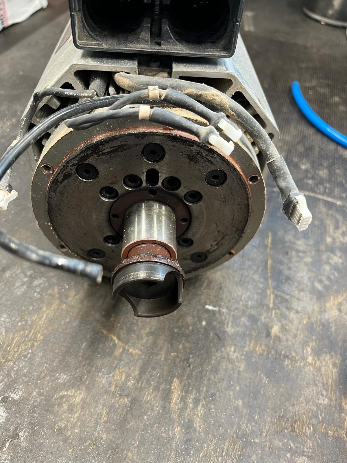 Zużyta głowica frezarska z widocznymi śladami eksploatacji i przewodami, leżąca na ciemnym, brudnym stole warsztatowym.