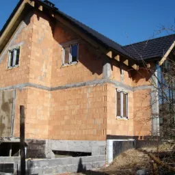 Dwukondygnacyjny dom murowany w trakcie budowy, widoczne ściany z czerwonej cegły, zamontowane okna PCV, czarna dachówka i izolacja fundamentów.