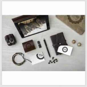 Kompozycja na jasnym drewnianym blacie: skórzane akcesoria, notes, długopis, pendrive, wizytówki z logo w stylu vintage, kulki i zdjęcie psa w ramce.