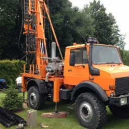 Pomarańczowy Unimog z wiertnicą na zielonym trawniku, obok składowane rury, dwóch mężczyzn w tle, krzewy i drzewa.