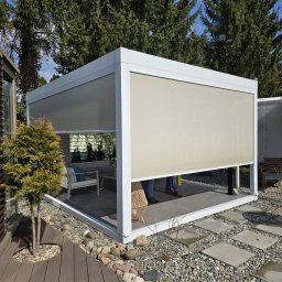 K-BRO sp. z o.o. - Nowoczesna, biała pergola z beżowymi osłonami przeciwsłonecznymi, ustawiona w ogrodzie z drewnianym tarasem i kamienną ścieżką, częściowo zasłonięta przez dwie osoby w jeansach.