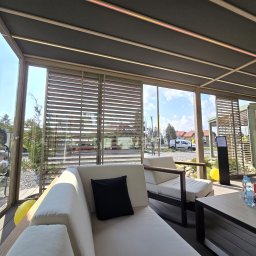 K-BRO sp. z o.o. - Zadaszona taras z żaluzjami zewnętrznymi, białymi sofami i drewnianym stołem, widok na ogród i zabudowania.