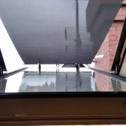 Widok z dołu na otwarte okno dachowe z zamontowaną, częściowo zasłoniętą, szarą tkaniną roletową; widoczny fragment muru z czerwonej cegły.