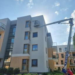 K-BRO sp. z o.o. - Montaż osłon okiennych na elewacji budynku wielorodzinnego z użyciem podnośnika koszowego, widoczny pracownik w koszu na tle błękitnego nieba.