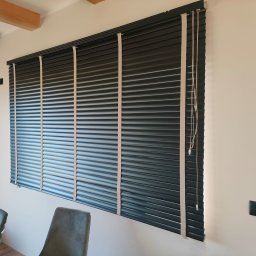 K-BRO sp. z o.o. - Ciemne drewniane żaluzje poziome z beżowymi taśmami na oknie w jasnym pokoju, widoczne fragmenty dwóch krzeseł i zabawki dziecięce.