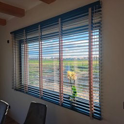 K-BRO sp. z o.o. - Żaluzje aluminiowe w kolorze szarym z drewnianymi akcentami, częściowo zasłonięte, z widokiem na krajobraz polny i kwiat w doniczce na parapecie.