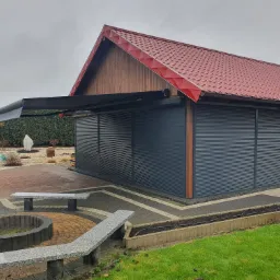 Zewnętrzny garaż z szarymi, zwiniętymi przesłonami, wysuwaną markizą i czerwonym dachem, otoczony brukowanym patio i zielonym trawnikiem w pochmurny dzień.