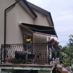 Czarna, rozłożona osłona przeciwsłoneczna nad balkonem domu z ozdobną, metalową balustradą i kwiatami w donicach.