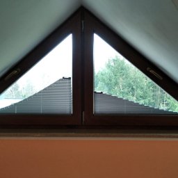 K-BRO sp. z o.o. - Trójkątne okno na poddaszu z drewnianą ramą i plisowanymi żaluzjami; widok na drzewa.