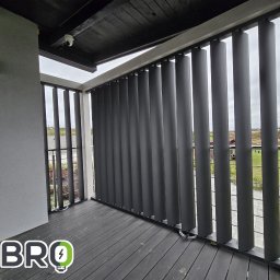 K-BRO sp. z o.o. - Szare pionowe osłony przeciwsłoneczne na balkonie z drewnianą podłogą kompozytową i widokiem na okolicę, zamontowana kamera monitoringu na suficie.