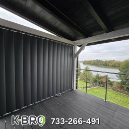 K-BRO sp. z o.o. - Ciemne, pionowe osłony okienne na balkonie z widokiem na rzekę, na drewnianej podłodze balkonu widoczne logo i numer telefonu wykonawcy.