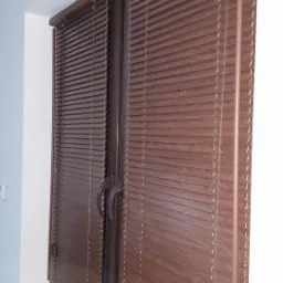 Zamknięte drewniane żaluzje poziome w kolorze orzechowym zamontowane w oknie dwuskrzydłowym, widoczne sznurki i uchwyty do regulacji. Parapet w kolorze zbliżonym do żaluzji.