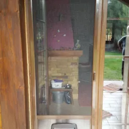 Drewniane drzwi balkonowe z moskitierą i zamontowanym przejściem dla zwierząt, widok na wnętrze z kominkiem i akcesoriami dla zwierząt.