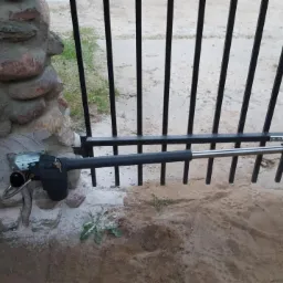 Automat do bramy wjazdowej zamontowany na kamiennym słupku i metalowej bramie, widoczne elementy mechaniczne i przewody.