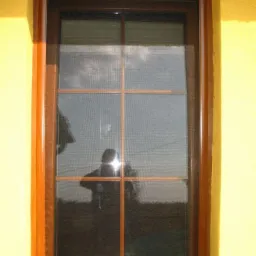 Okno z brązową ramą i siatką, widoczny refleks sylwetki fotografa, żółta ściana budynku w tle.