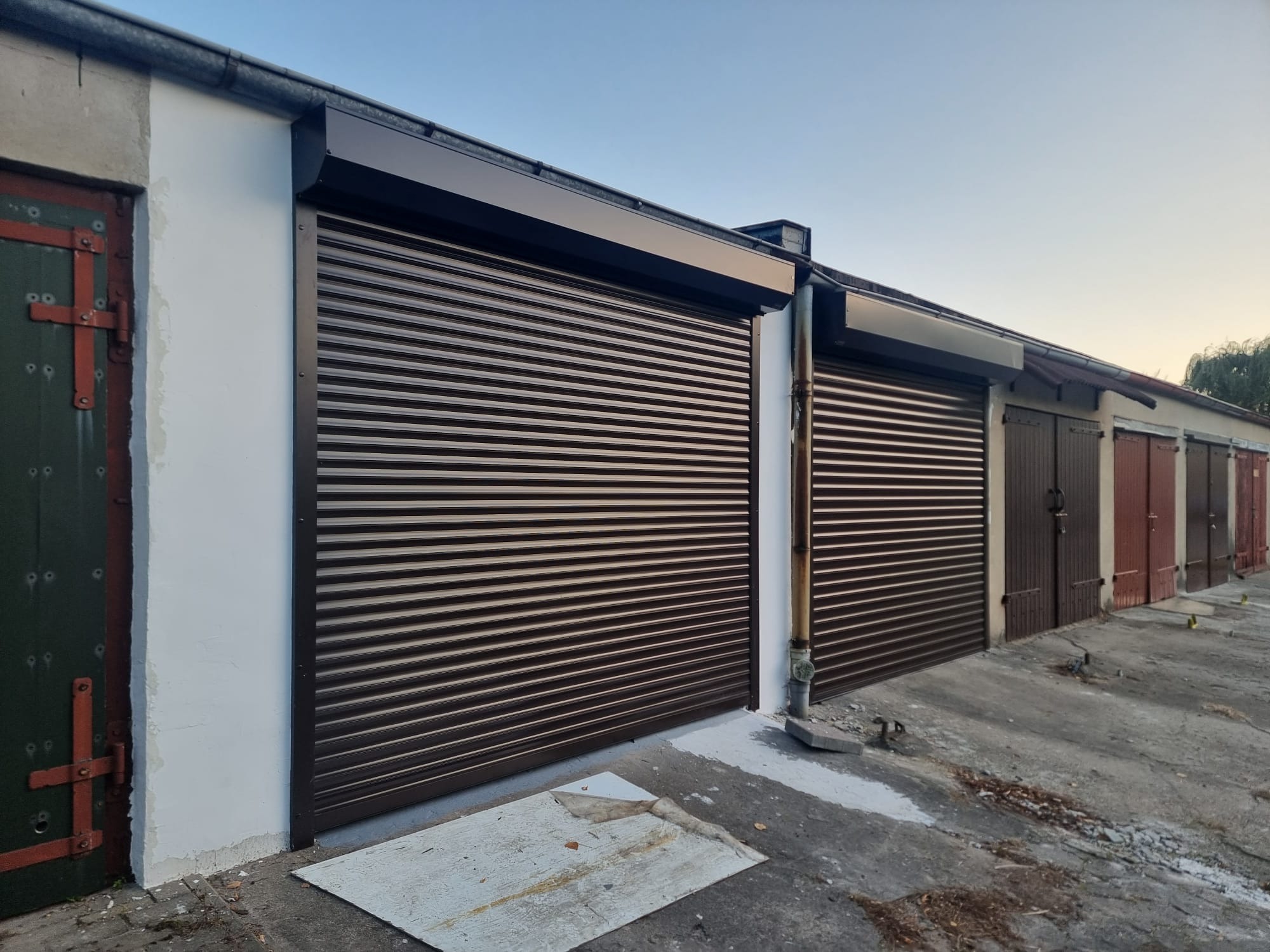 Zewnętrzne, brązowe osłony okienne typu rolowanego na garażach, widoczne ślady niedawnego malowania na posadzce i ścianie.