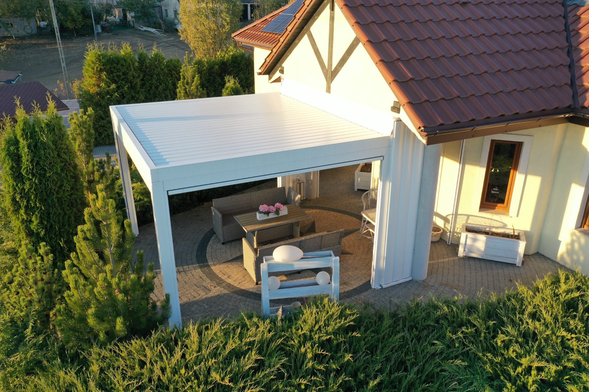 Nowoczesna, biała pergola tarasowa z meblami wypoczynkowymi i dekoracjami, widok z góry na tle domu z brązowym dachem i zieleni ogrodu.