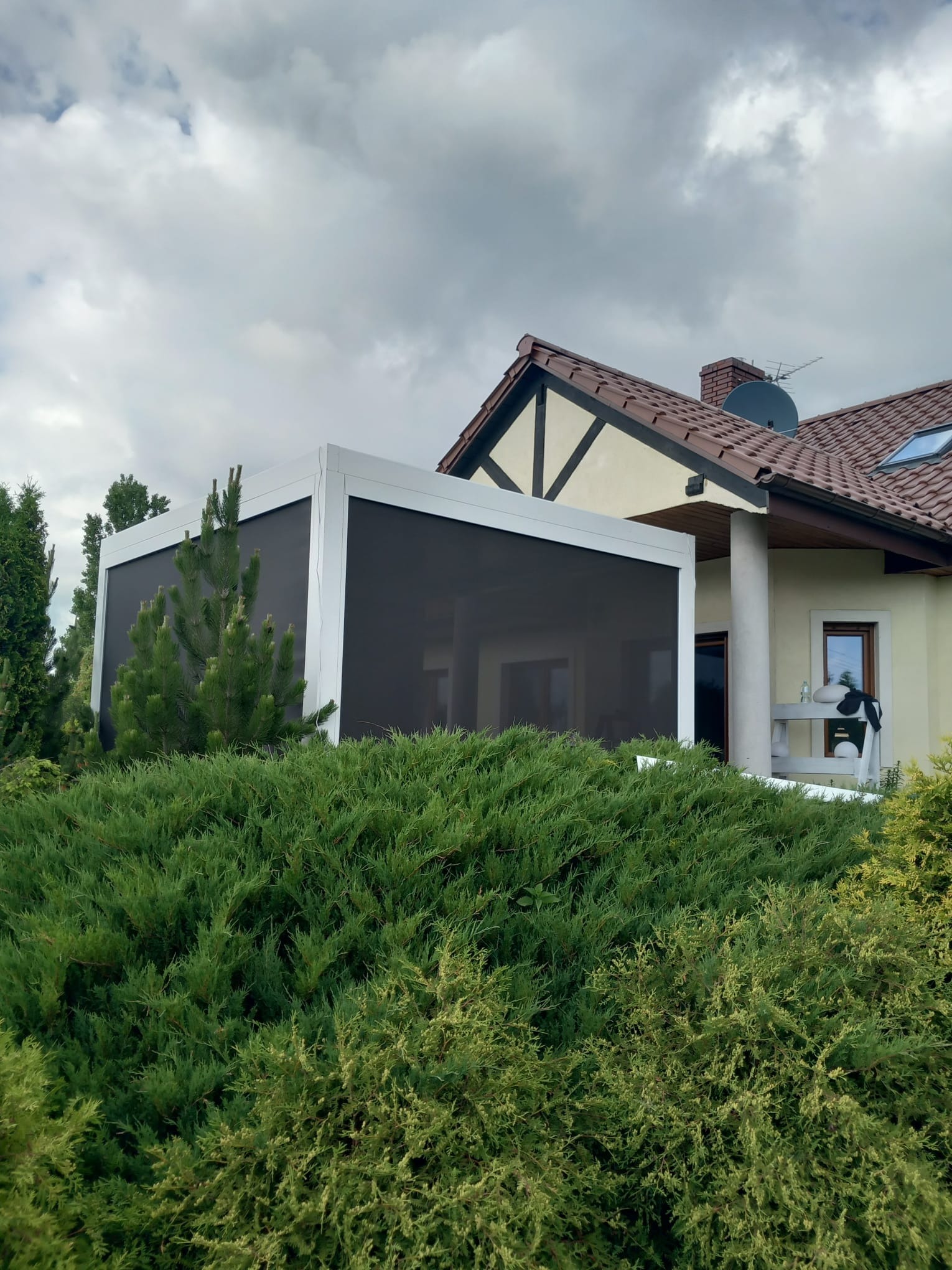 Nowoczesna pergola z białymi ramami i zaciemniającą przesłoną, przylegająca do domu z beżową elewacją i brązowym dachem, otoczona zielonymi krzewami.