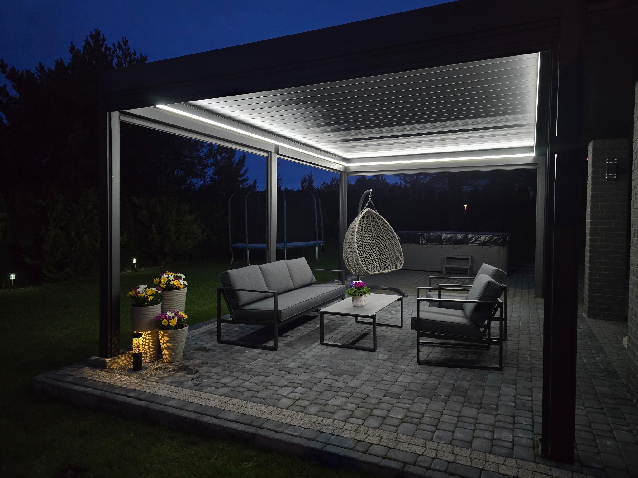 Nowoczesna pergola tarasowa z oświetleniem LED wbudowanym w lamele, szara sofa i fotele ogrodowe na brukowanym patio, wiszące krzesło i jacuzzi w tle, nocne oświetlenie ogrodu.