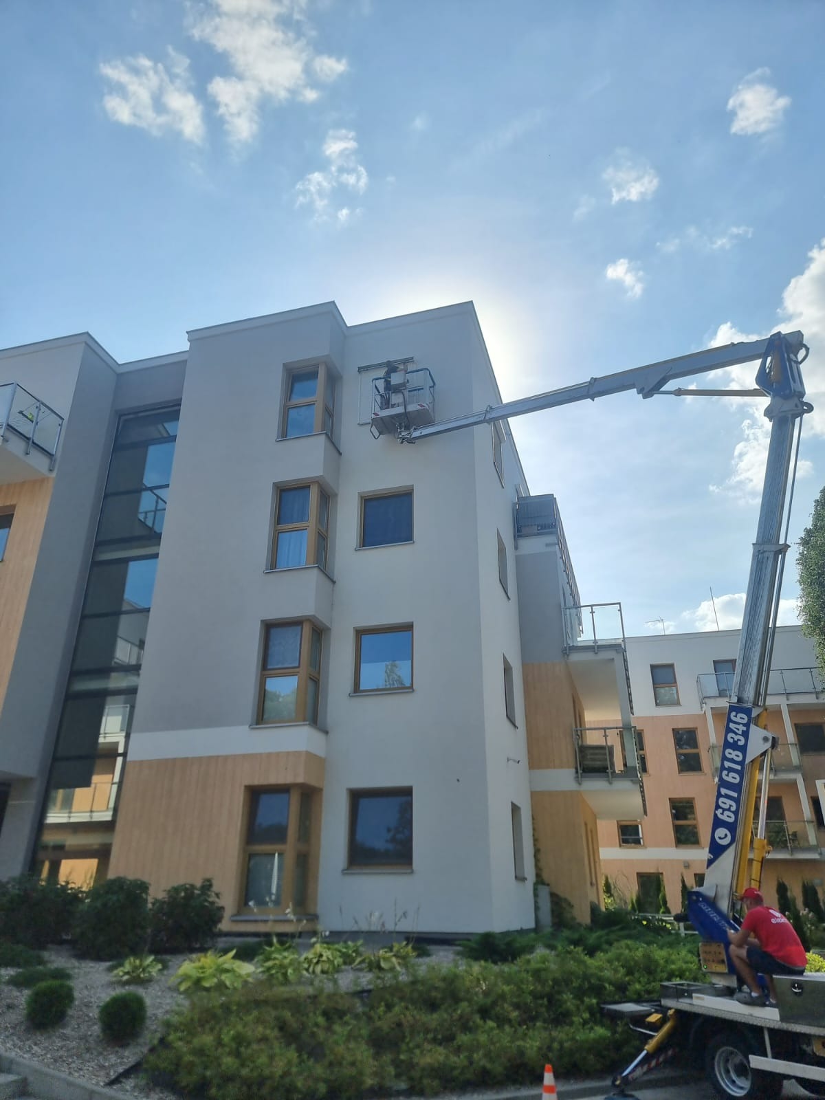 Montaż osłon okiennych na elewacji budynku wielorodzinnego z użyciem podnośnika koszowego, widoczny pracownik w koszu na tle błękitnego nieba.