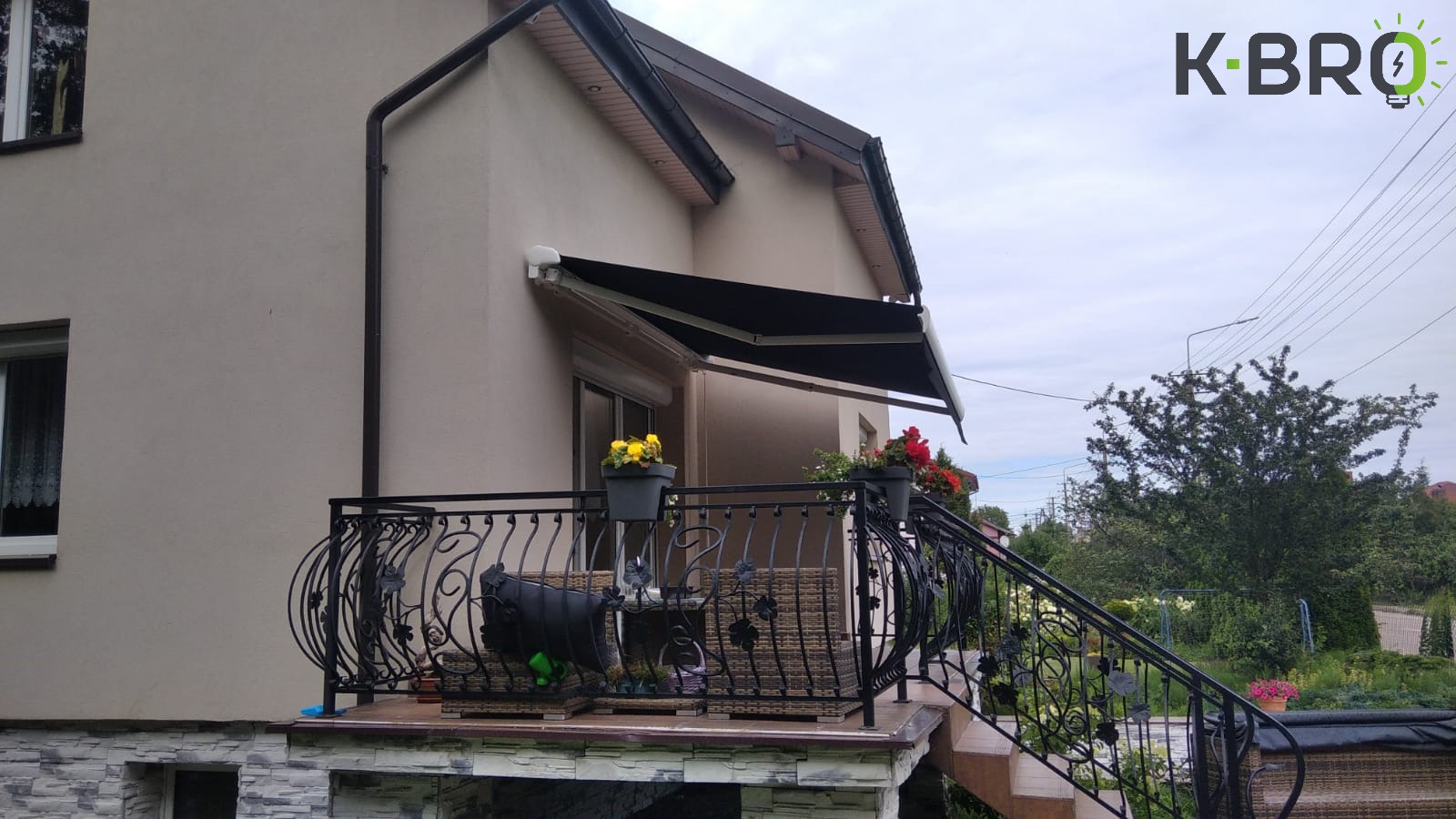 Czarna rozkładana osłona przeciwsłoneczna nad balkonem z kutą balustradą, widok z dołu na dwukondygnacyjny dom z jasną elewacją i logo firmy w prawym górnym rogu.