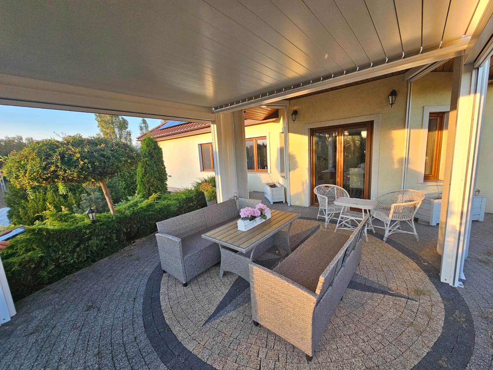 Zadaszony taras z meblami ogrodowymi z technorattanu i widokiem na zadbany ogród w ciepłym, popołudniowym świetle.