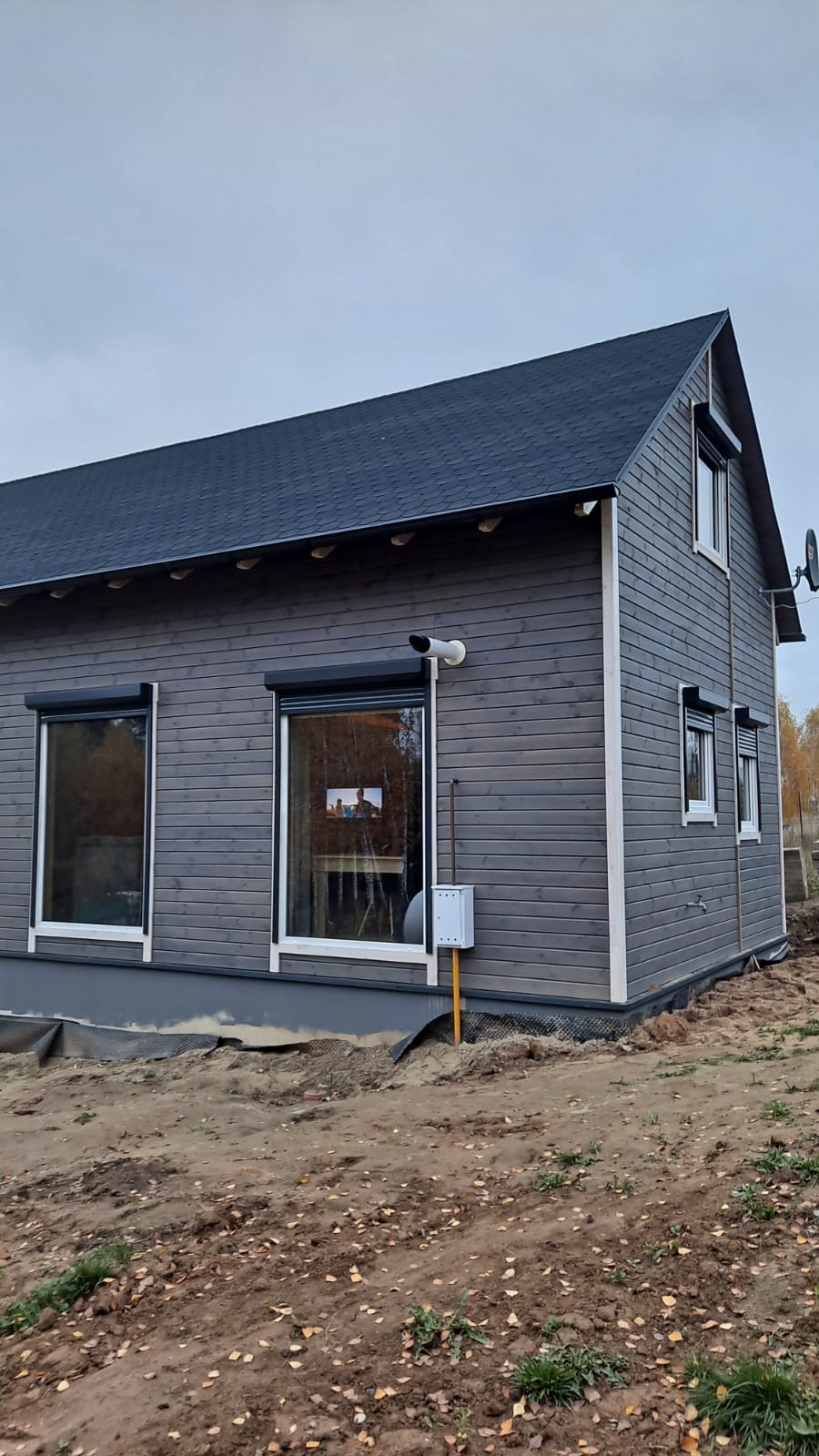 Szary dom z drewnianą elewacją, częściowo opuszczone czarne rolety okienne, instalacja gazowa przy ścianie, antena satelitarna na dachu
