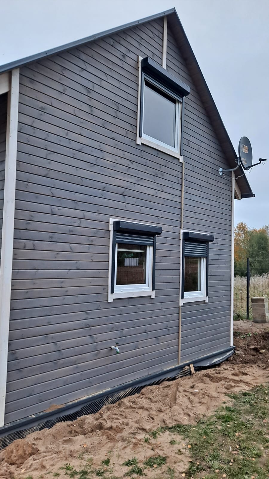 Szary drewniany dom z trzema oknami zabezpieczonymi czarnymi roletami zewnętrznymi, widoczna antena satelitarna i teren wokół domu w trakcie zagospodarowania.