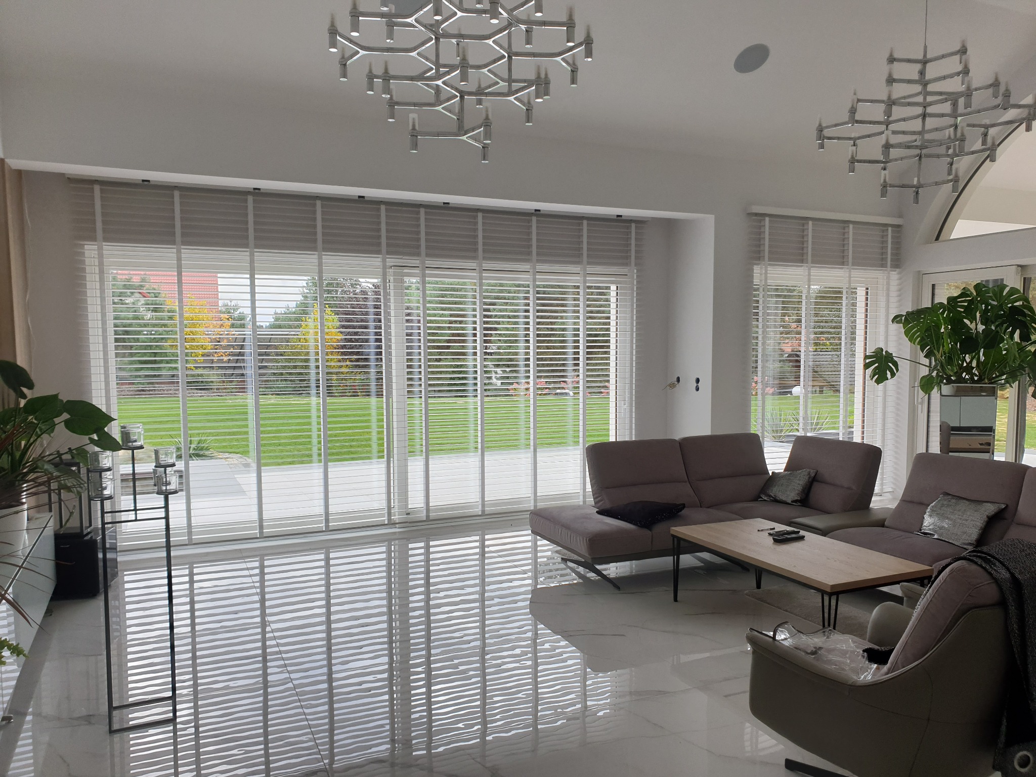 Jasne, przestronne wnętrze z dużymi oknami przesłoniętymi poziomymi osłonami okiennymi, odbijającymi światło na błyszczącej podłodze.