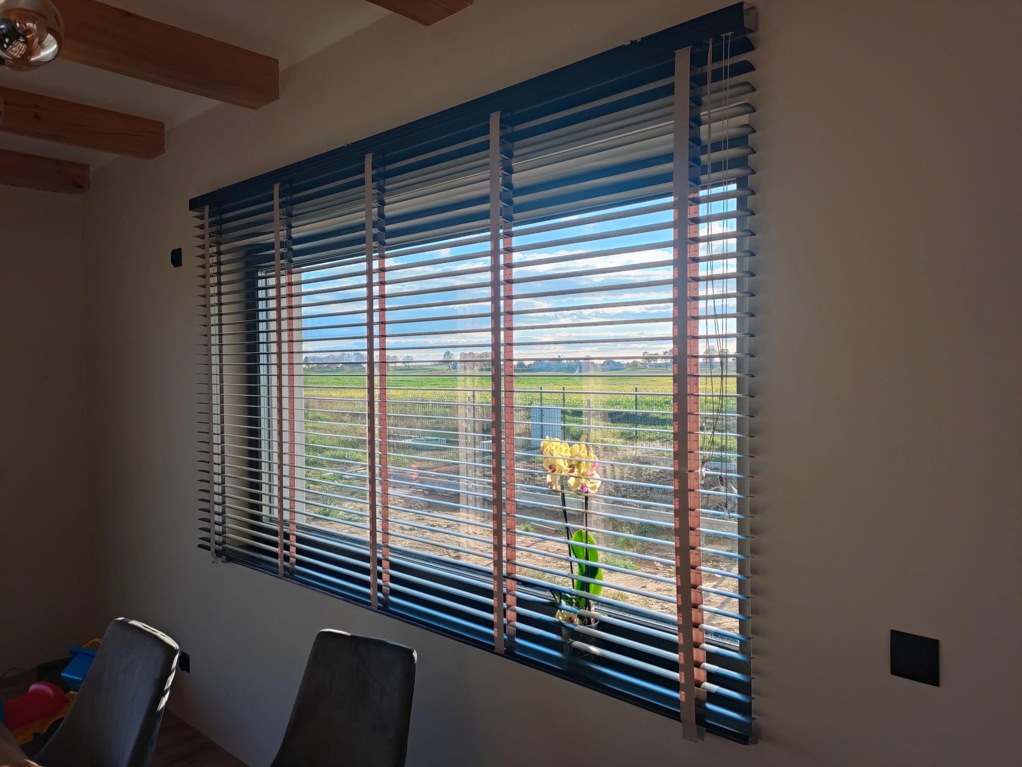 Żaluzje aluminiowe w kolorze szarym z drewnianymi akcentami, częściowo zasłonięte, z widokiem na krajobraz polny i kwiat w doniczce na parapecie.