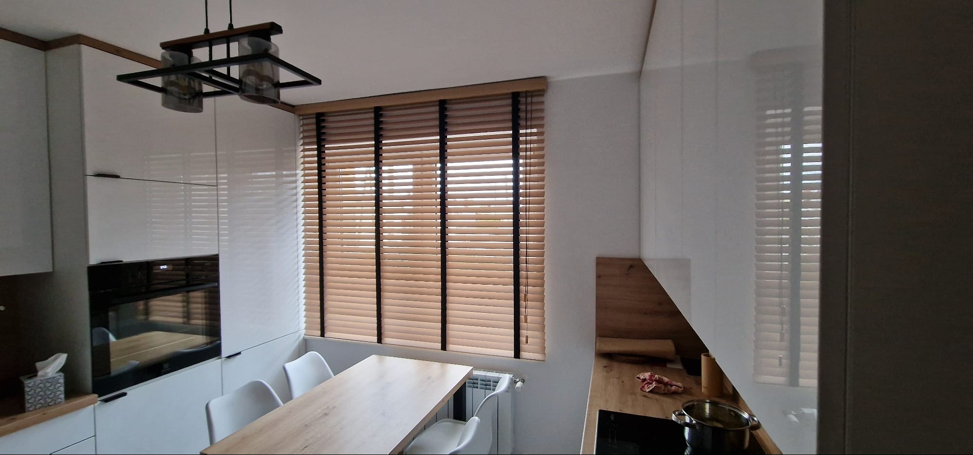 Drewniane poziome listwy okienne z czarnymi sznurkami w nowoczesnej kuchni z białymi szafkami i drewnianym blatem stołu.
