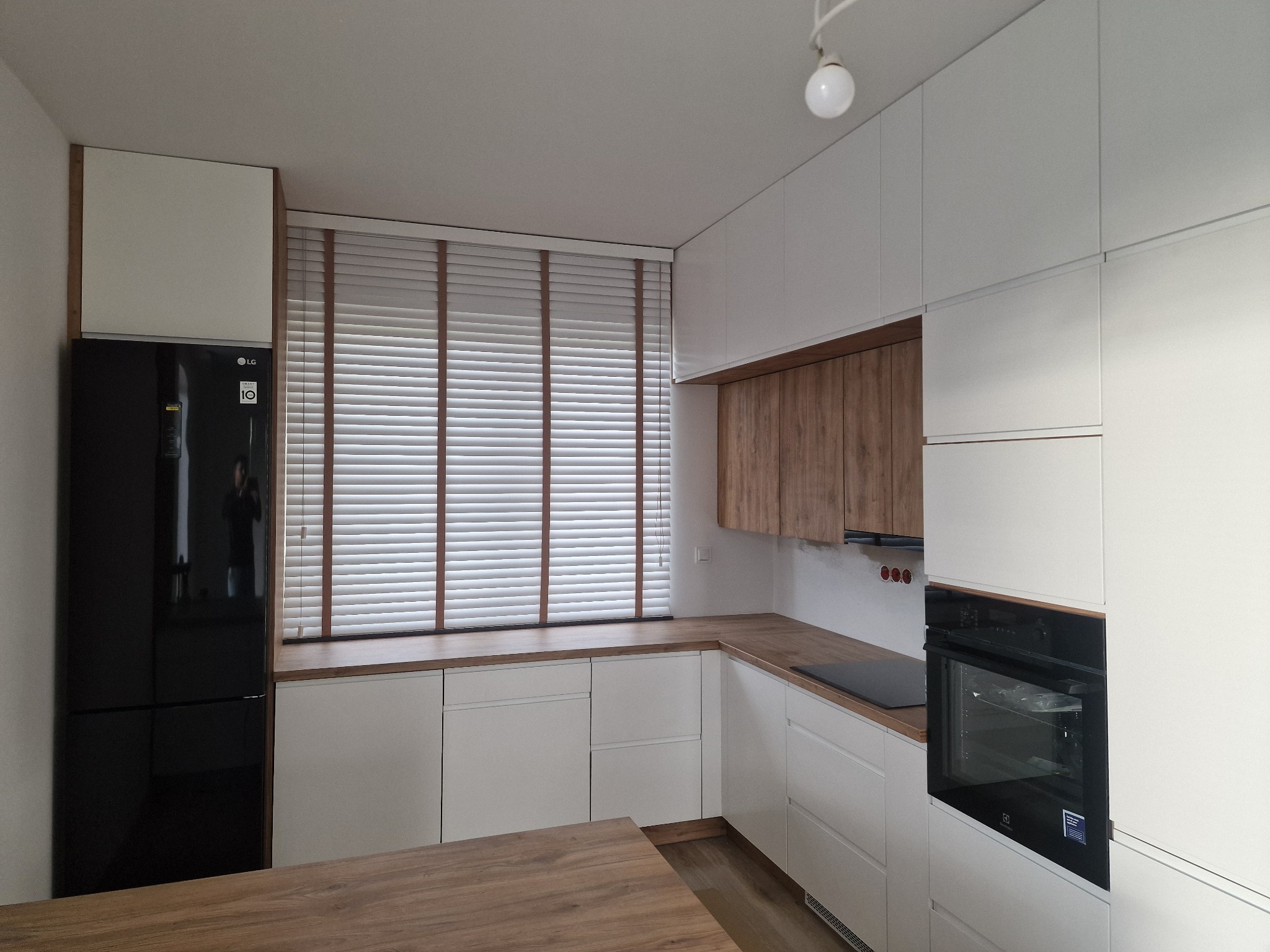 Nowoczesna kuchnia z białymi szafkami, drewnianymi blatami i żaluzjami poziomymi z drewnianymi lamelami, widok na czarny piekarnik i lodówkę.