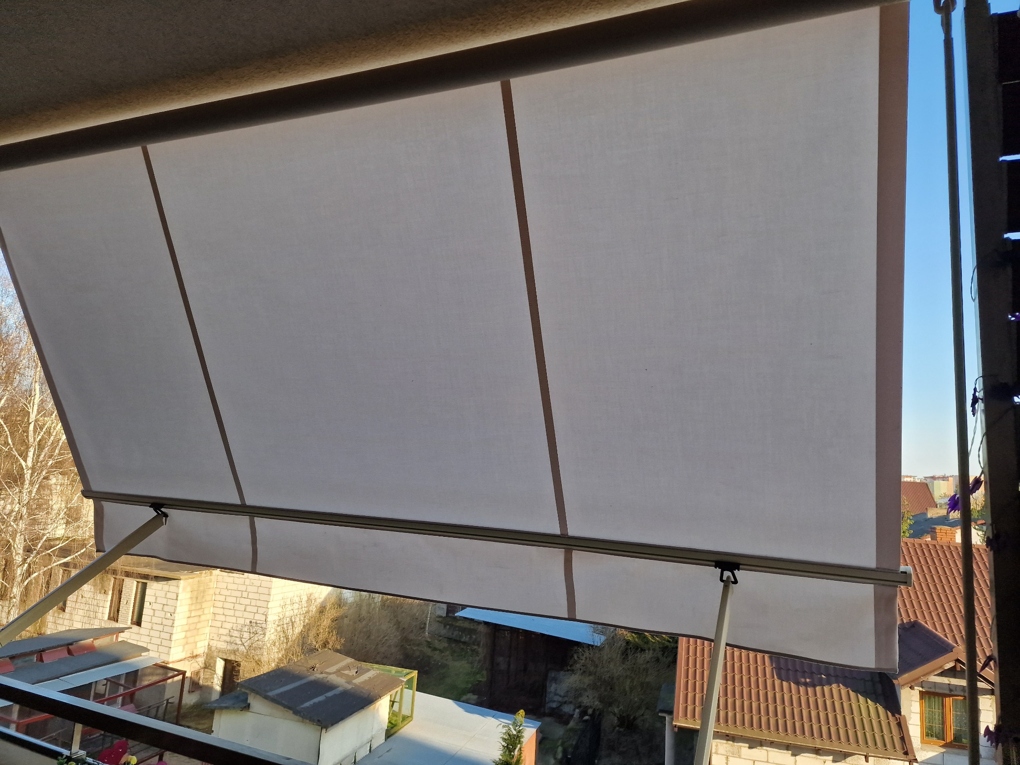 Zasłonięty balkon tkaniną w jasnym kolorze, widok z góry na zabudowania i drzewa w słoneczny dzień.