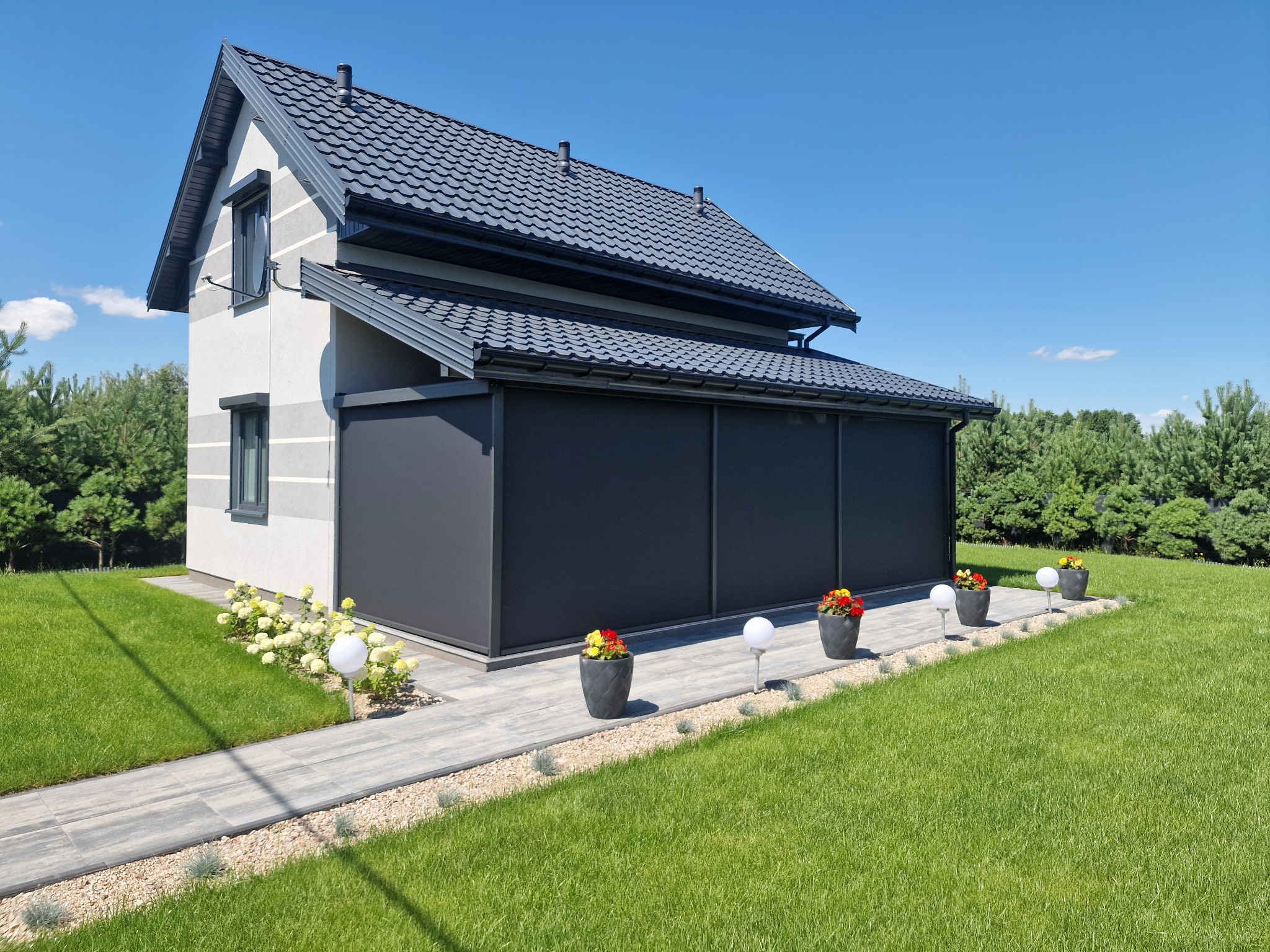 Nowoczesny dom z szarymi roletami zewnętrznymi, oświetlonym chodnikiem i zadbanym trawnikiem w słoneczny dzień.