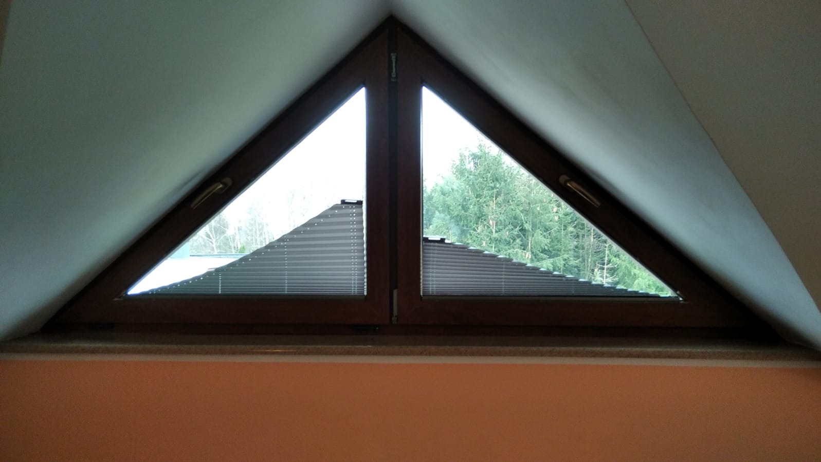 Trójkątne okno na poddaszu z drewnianą ramą i plisowanymi żaluzjami; widok na drzewa.