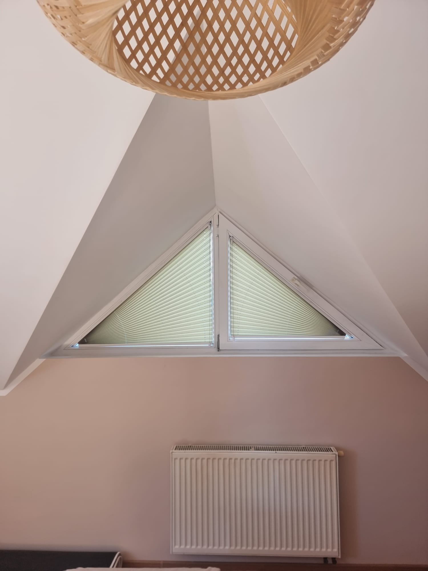 Okno na poddaszu o trójkątnym kształcie z zamontowanymi plisowanymi zasłonami w kolorze jasnej zieleni, widoczny grzejnik i dekoracyjna lampa sufitowa z plecionki.
