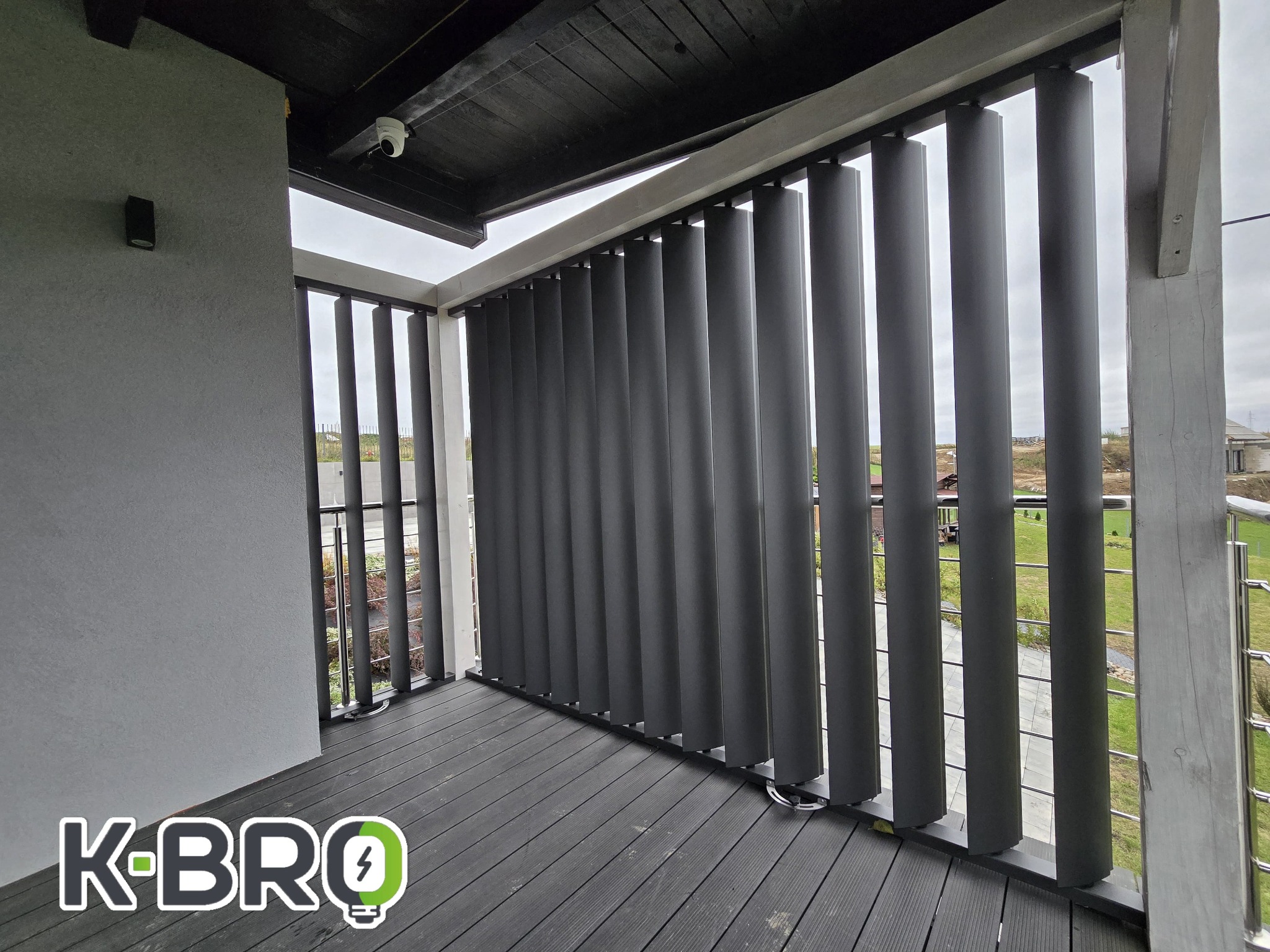 Szare pionowe osłony przeciwsłoneczne na balkonie z drewnianą podłogą kompozytową i widokiem na okolicę, zamontowana kamera monitoringu na suficie.