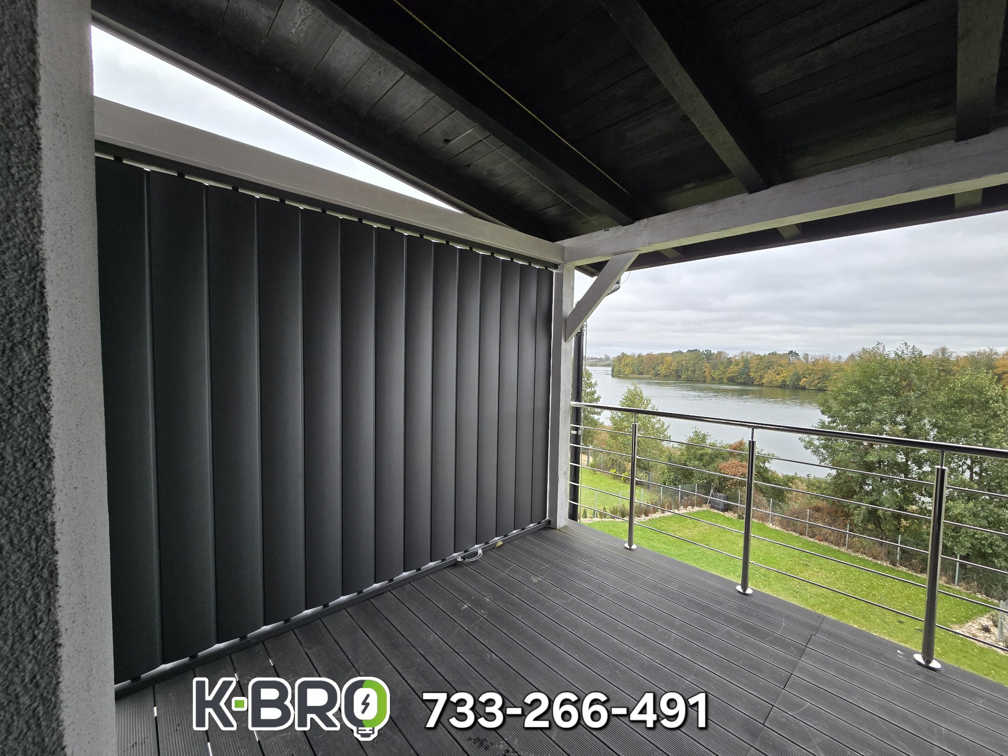 Ciemne, pionowe osłony okienne na balkonie z widokiem na rzekę, na drewnianej podłodze balkonu widoczne logo i numer telefonu wykonawcy.